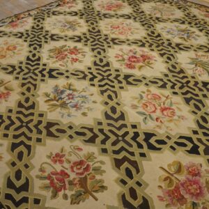 Low-pile beige rug with dark geometric motifs outlined in olive, framing multi-colored floral clusters on plank flooring.