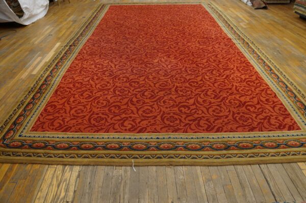 Expansive orange rug with dense red scrollwork pattern and complex gold, blue, and olive borders on wood flooring.