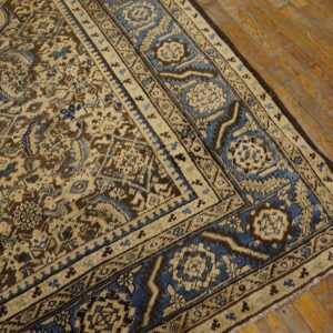 Intricate traditional rug pattern featuring brown, tan, and blue colors, shown on warm wood flooring.