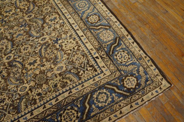 Intricate traditional rug pattern featuring brown, tan, and blue colors, shown on warm wood flooring.