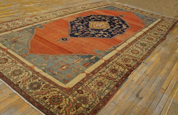 Orange and navy medallion rug with detailed tan and blue borders on rustic, light wooden flooring.