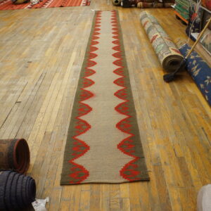 Tan flatweave runner with red zigzag borders sits on a light wood floor near many stacked rolled rugs.