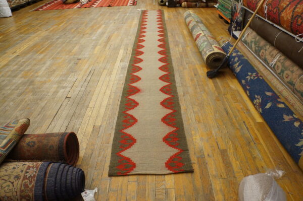 Tan flatweave runner with red zigzag borders sits on a light wood floor near many stacked rolled rugs.