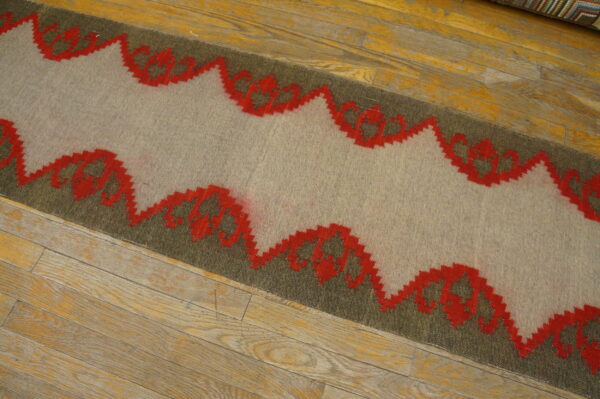 Gray and olive flatweave rug with a red zig-zag geometric pattern lies on worn hardwood planks.