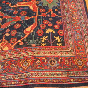 Ornate patterned rug in navy blue, red, and gold, showing borders and field details on light wood flooring.