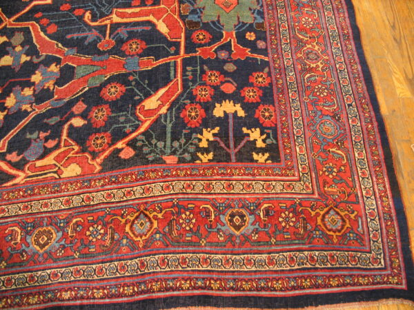 Ornate patterned rug in navy blue, red, and gold, showing borders and field details on light wood flooring.