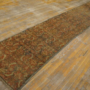 Muted red and olive patterned runner rug lying lengthwise on worn, yellow-toned wooden planks.