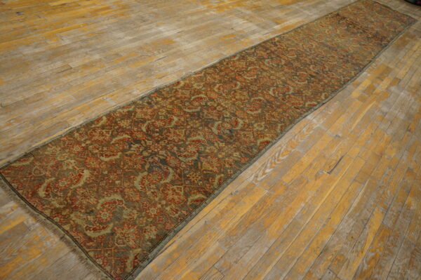 Muted red and olive patterned runner rug lying lengthwise on worn, yellow-toned wooden planks.