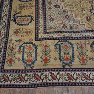 Tan rug corner with stylized floral boteh motifs in blue, red, and green on rustic wood planks.