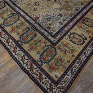 Muted tan rug corner with complex blue and red floral borders sits on distressed light wood flooring.
