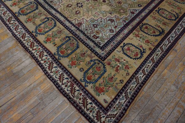 Muted tan rug corner with complex blue and red floral borders sits on distressed light wood flooring.