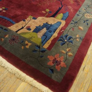 Close-up of a burgundy scenic rug with a gray floral border and fringe on distressed wood flooring.