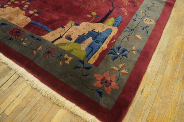 Close-up of a burgundy scenic rug with a gray floral border and fringe on distressed wood flooring.