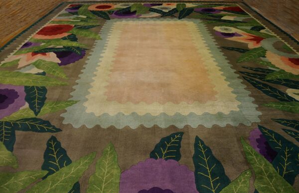 Oversized rug with a bold, colorful floral border surrounding a light peach scalloped central field on wood flooring.