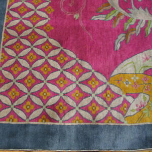 Pink rug corner with geometric trellis and stylized floral motifs framed by a wide blue border on wood floors.