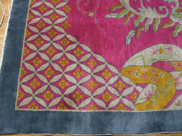 Pink rug corner with geometric trellis and stylized floral motifs framed by a wide blue border on wood floors.