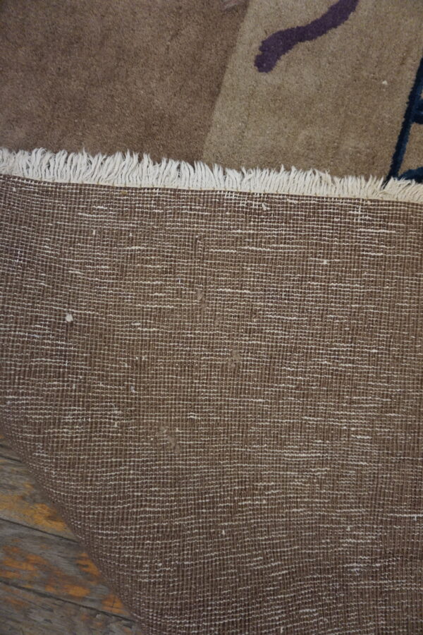 View showing the woven brown backing, cream fringe, and tan pile with dark purple accents on wood plank flooring.