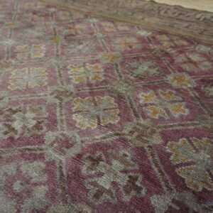 Textured close-up of a purple rug with geometric grey and gold repeating medallion motifs and a brown border.