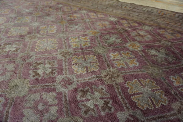 Textured close-up of a purple rug with geometric grey and gold repeating medallion motifs and a brown border.