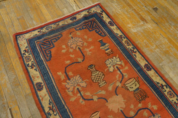 Rust orange and blue rug with floral and vase patterns lies on distressed yellow-toned hardwood floor.