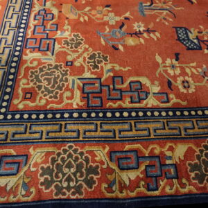 Reddish-orange rug field with blue floral patterns and a wide border of geometric Greek key designs.