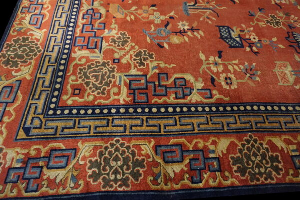 Reddish-orange rug field with blue floral patterns and a wide border of geometric greek key designs.