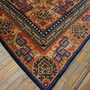 Detailed corner of a red, navy, and gold rug displaying key and floral patterns on worn wood.