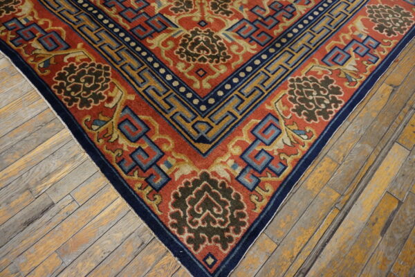 Detailed corner of a red, navy, and gold rug displaying key and floral patterns on worn wood.