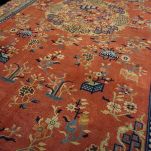 Red-orange rug featuring detailed blue and yellow botanical motifs and a dark geometric meander border.