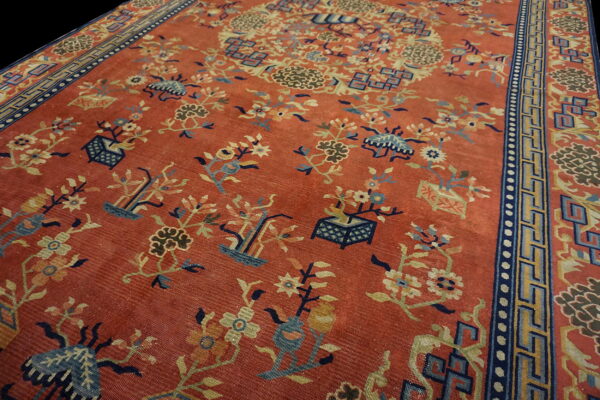 Red-orange rug featuring detailed blue and yellow botanical motifs and a dark geometric meander border.