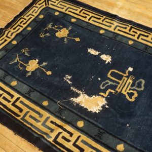 Dark blue rug with yellow geometric border and floral designs shows large holes exposing the wood floor.