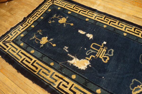 Dark blue rug with yellow geometric border and floral designs shows large holes exposing the wood floor.