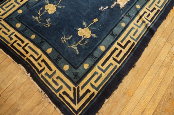 Low-pile indigo rug with a gold floral field and wide geometric key border on light wood flooring.