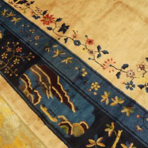 Close-up of a tan rug with a navy and teal border featuring floral and landscape designs on distressed yellow wood flooring.