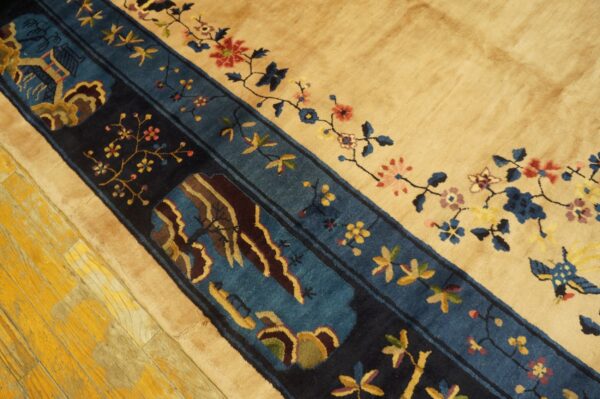 Close-up of a tan rug with a navy and teal border featuring floral and landscape designs on distressed yellow wood flooring.