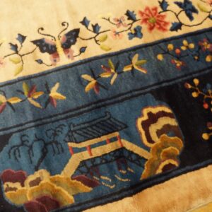 Detailed view of a cream rug field and dark blue border featuring floral, butterfly, and pagoda motifs on a wood floor.