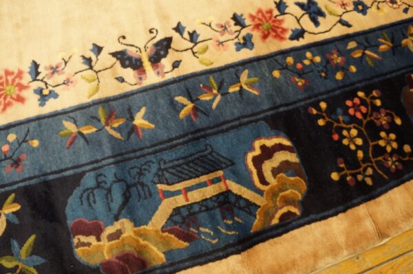 Detailed view of a cream rug field and dark blue border featuring floral, butterfly, and pagoda motifs on a wood floor.