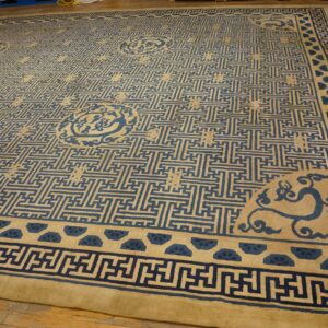 Tan rug with a dense blue interlocking maze pattern and ornamental borders rests on hardwood flooring.