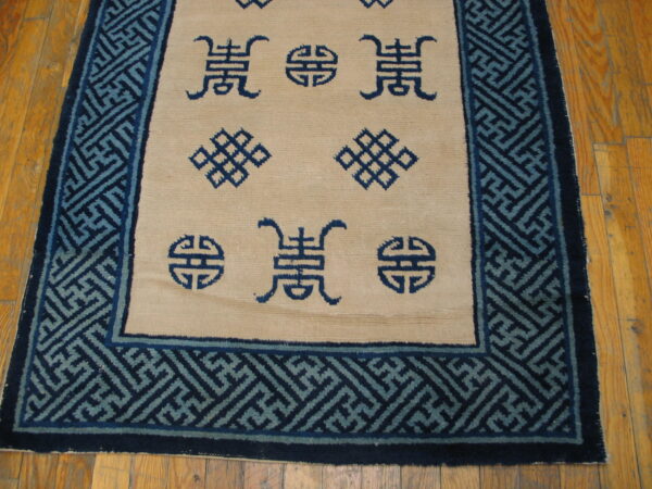 Tan rug with dark blue symbols and a wide key pattern border rests on visible hardwood flooring.