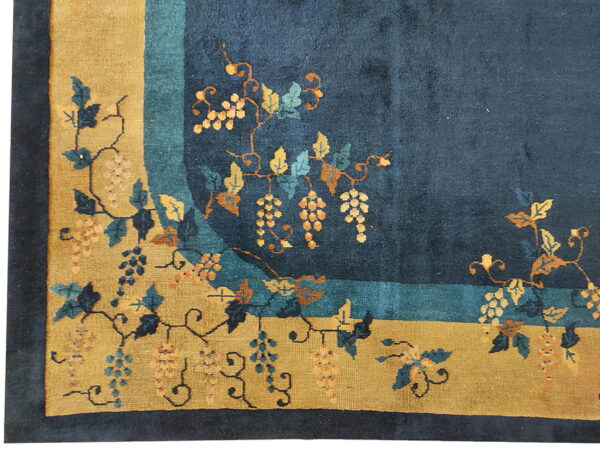 Corner view of a high-pile rug showing a navy field, gold border, and stylized grape and vine pattern.