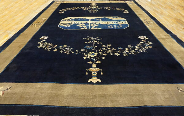 Large navy blue rug with a gold border features floral vines and a blue landscape cartouche on wood flooring.