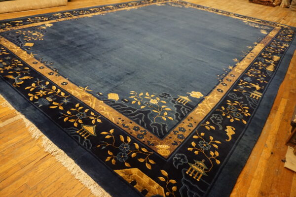 Deep blue rug featuring a wide scenic border with gold floral motifs, birds, and pagodas on wood plank flooring.