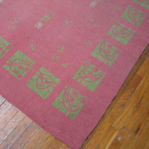 Pink, low-pile rug featuring a border of green squares with stylized foliate patterns on wood floor.