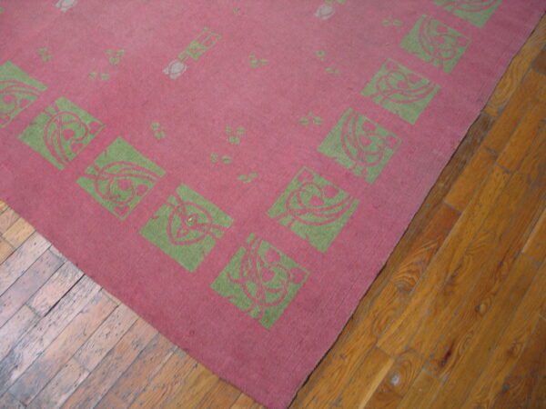Pink, low-pile rug featuring a border of green squares with stylized foliate patterns on wood floor.