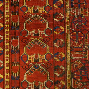 Textured close-up showing a red rug with complex geometric patterns in blue, yellow, and cream on wood.