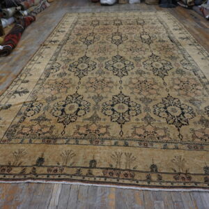 Tan low-pile rug with geometric floral motifs rests on worn wood flooring near rolled rugs.