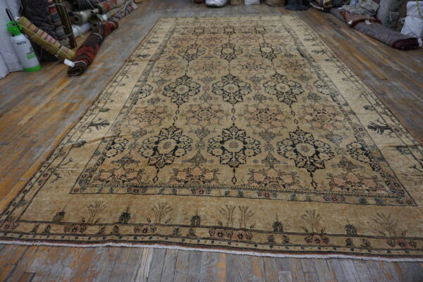 Tan low-pile rug with geometric floral motifs rests on worn wood flooring near rolled rugs.