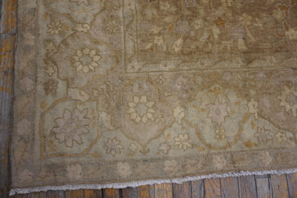 Muted beige and tan traditional rug with a detailed floral pattern and fringe on rustic wood flooring.