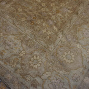 Textured rug corner in muted beige and brown with floral patterns, placed on worn wood flooring.