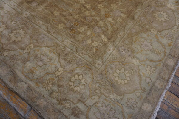 Textured rug corner in muted beige and brown with floral patterns, placed on worn wood flooring.